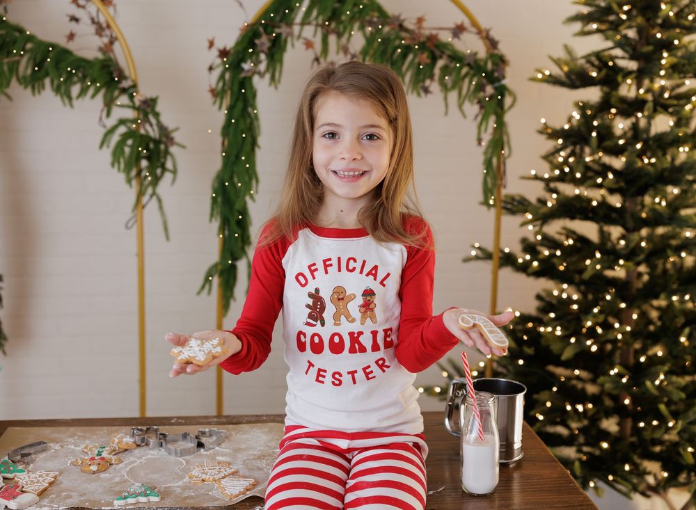 Girl in Christmas pajamas and Official Cookie Tester shirt holds cookies near a decorated tree.