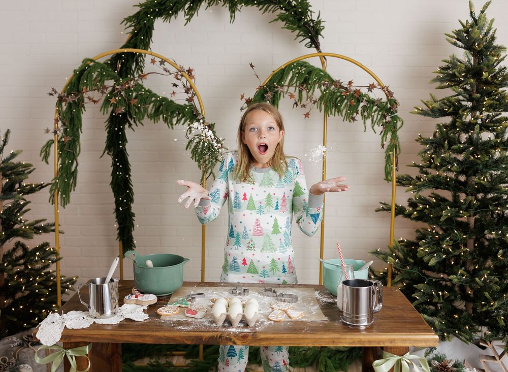 Girl in Christmas pajamas baking cookies at a wooden table, surrounded by holiday trees and festive garland arch.