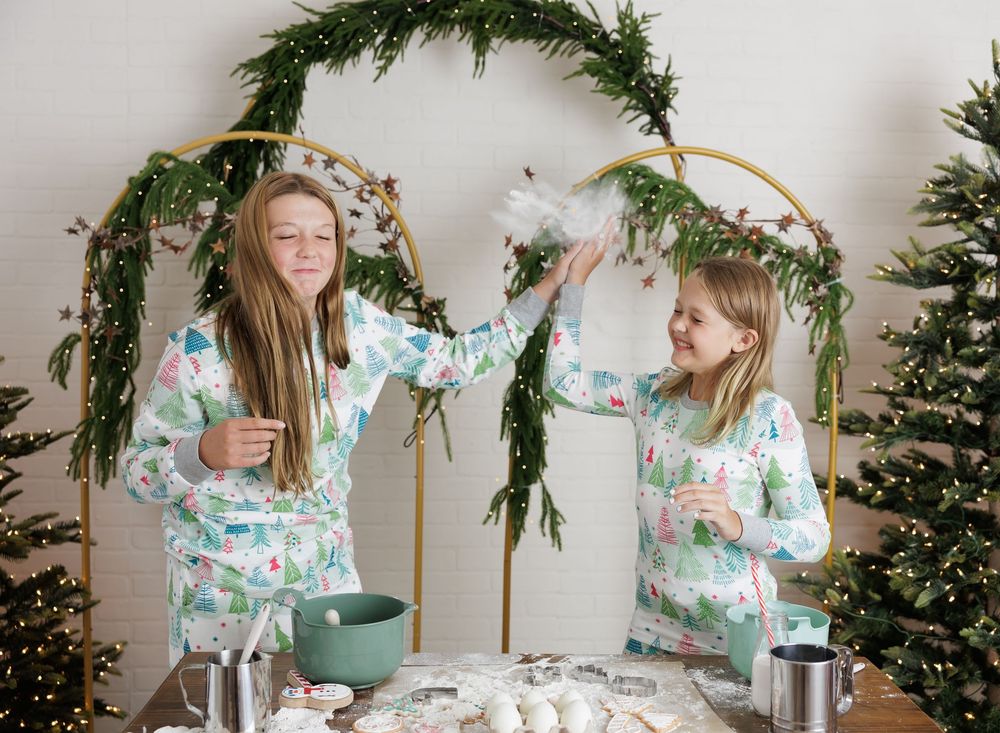 Two girls in matching floral pajamas decorating a Christmas arch with greenery near holiday trees.