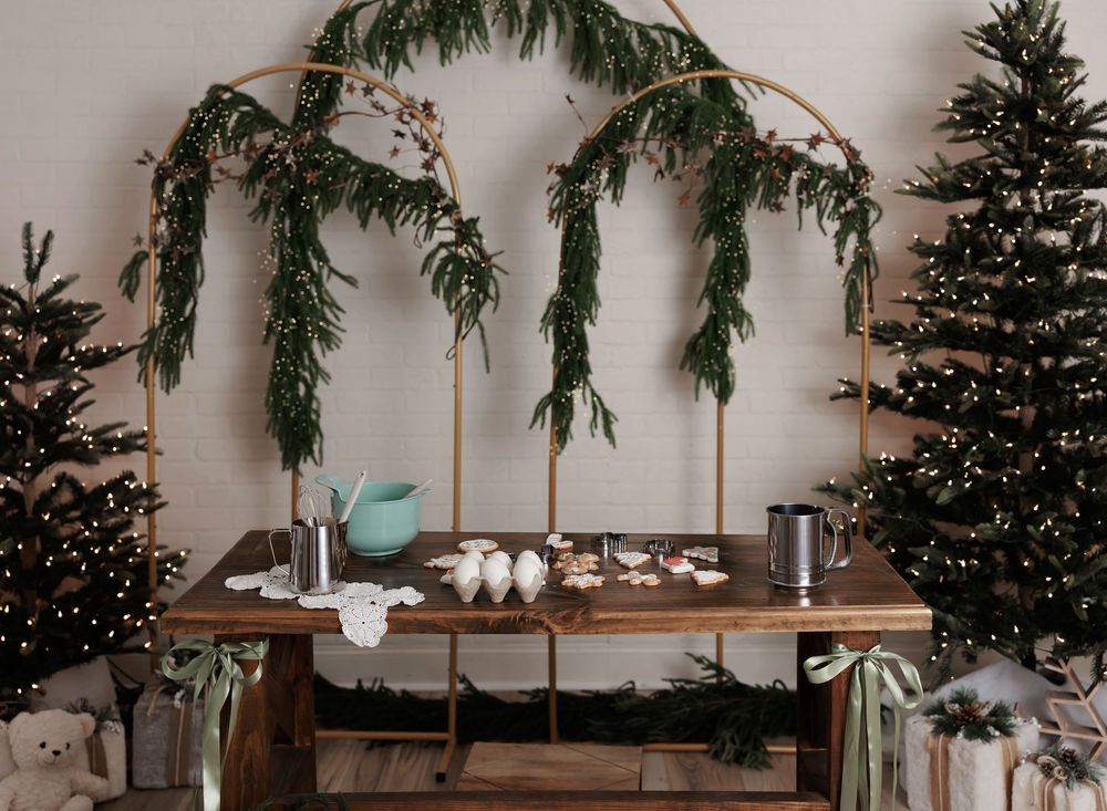 Gold arch with greenery over a rustic wooden table decorated for Christmas, flanked by lit Christmas trees.