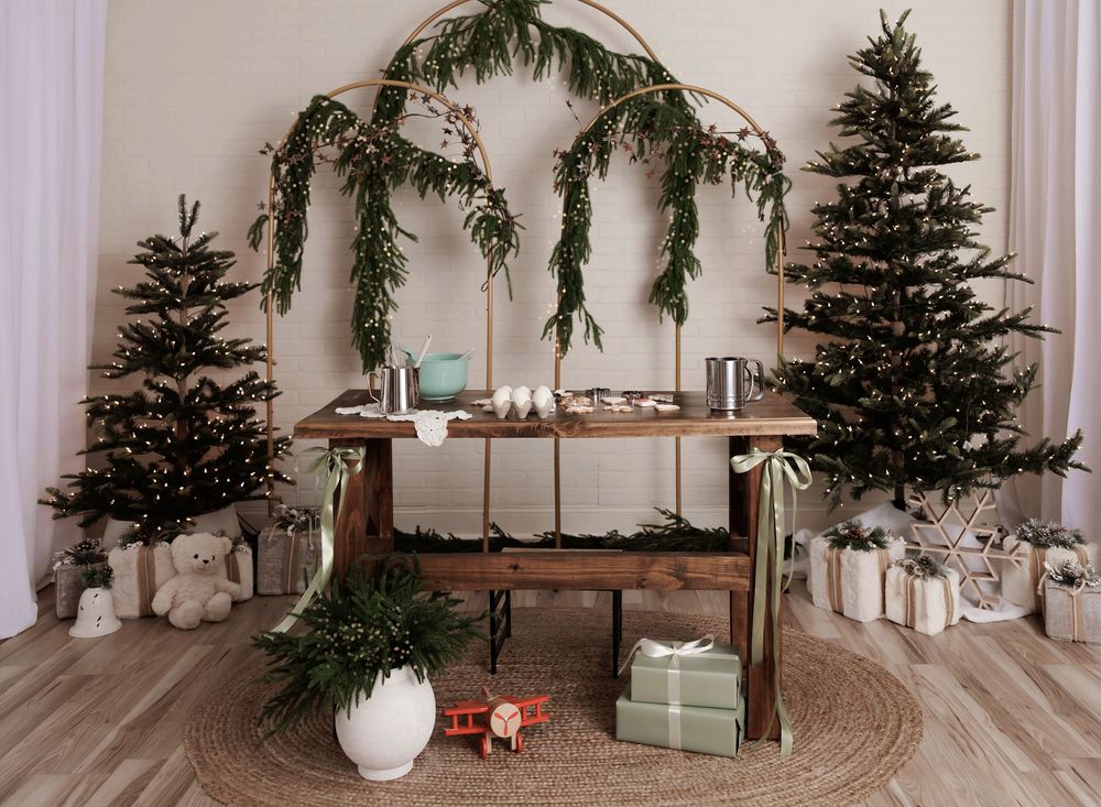 Festive Christmas vignette with decorated trees, wrapped gifts, wooden table, and garland arch on white backdrop.