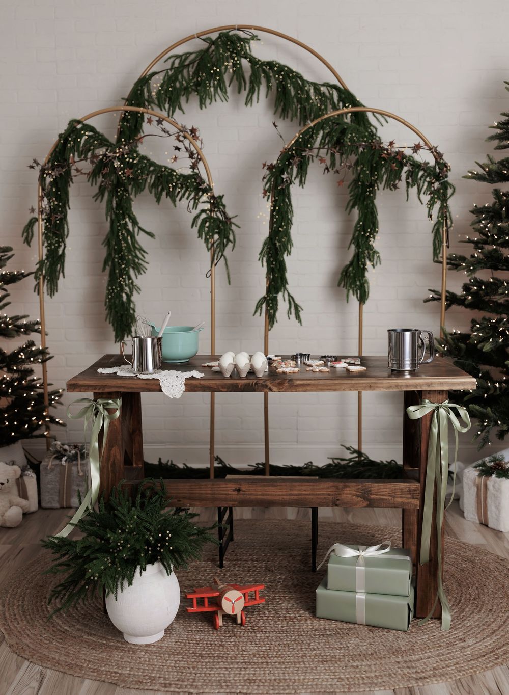 Rustic wooden table with holiday greenery arch, Christmas trees, and wrapped gifts in a festive winter decor setting.