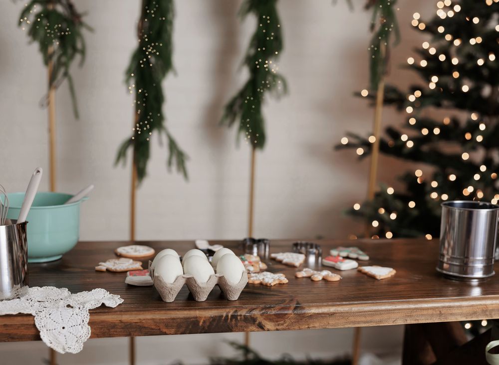 Rustic wooden table with eggs, Christmas cookies, and cookie cutters near a decorated tree with warm lights.