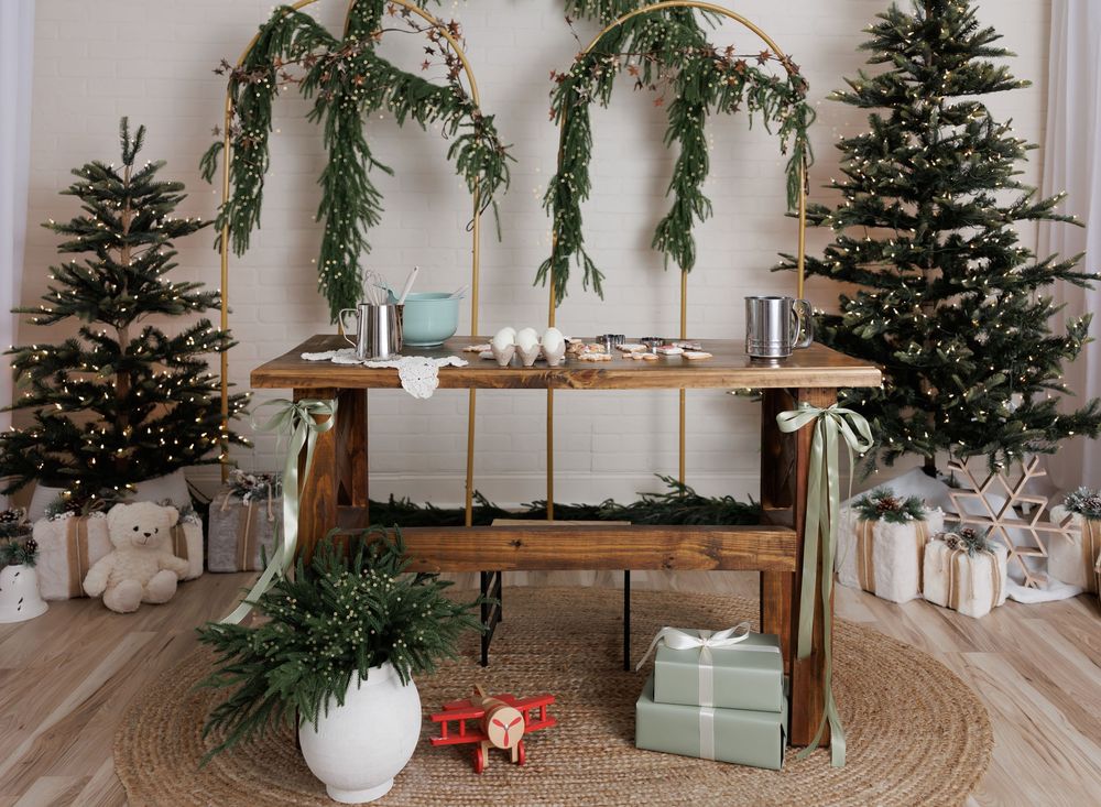 Rustic wooden table with holiday greenery arch, flanked by decorated Christmas trees and wrapped gifts.
