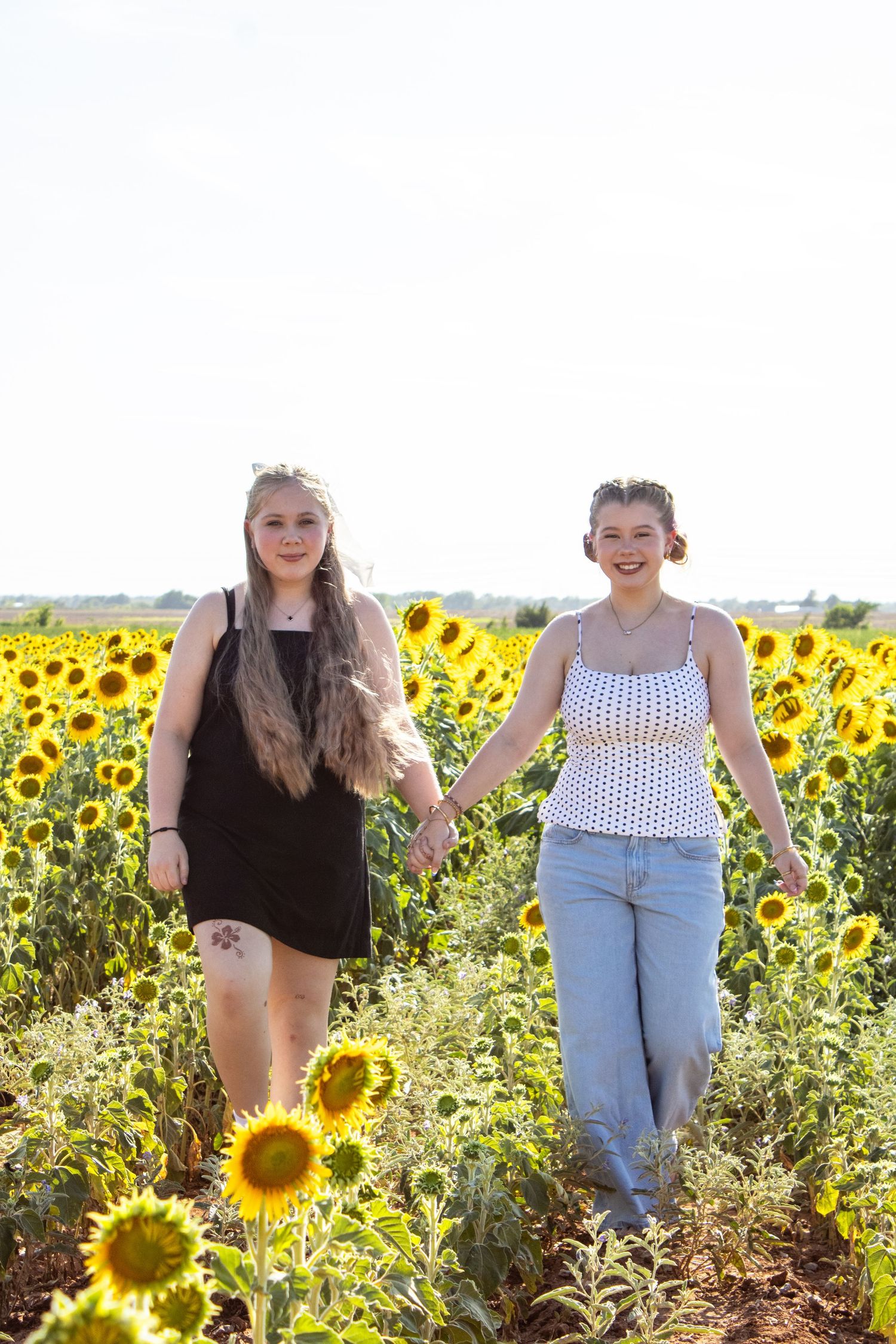 Two young women hold hands while walking through a bright sunny sunflower field together.