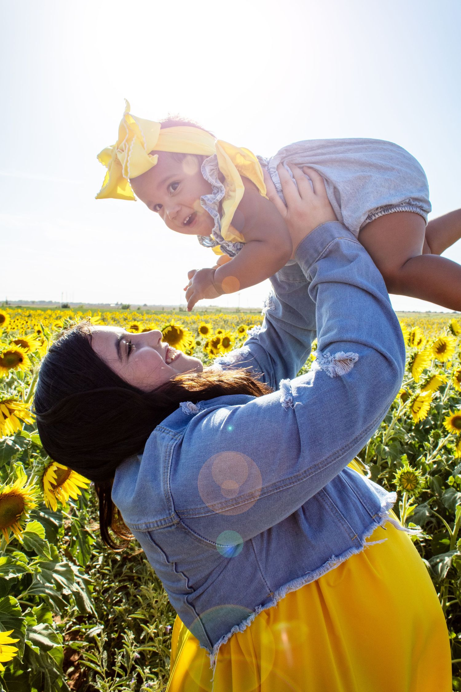 Mom holding baby with yellow bow overhead in sunny sunflower field.