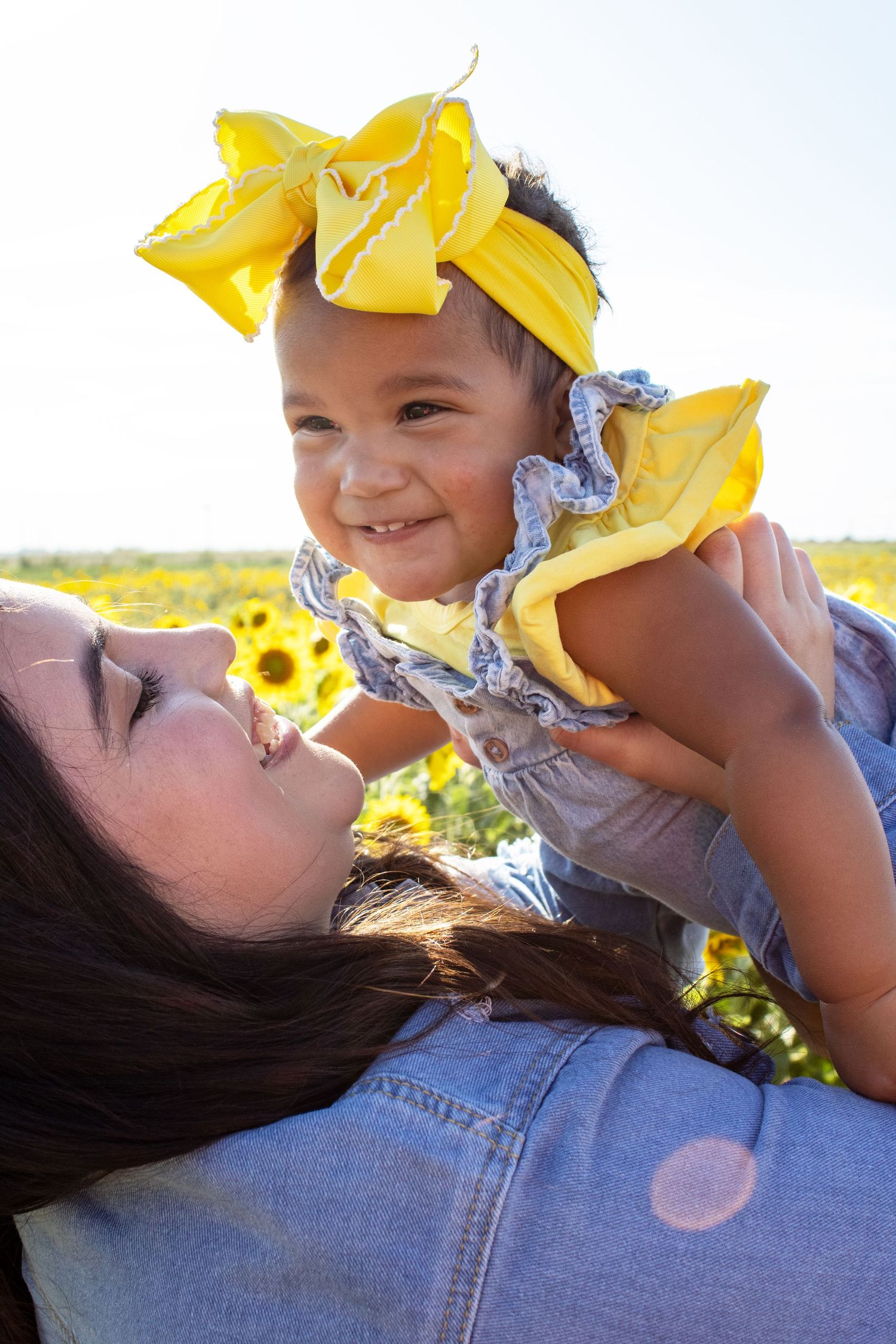 Mother lifting smiling baby girl with yellow bow in sunflower field.