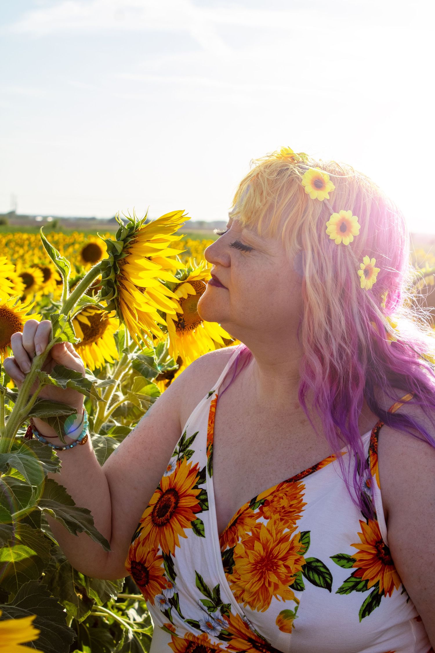 Woman with purple hair smelling sunflower in bright sunny field.