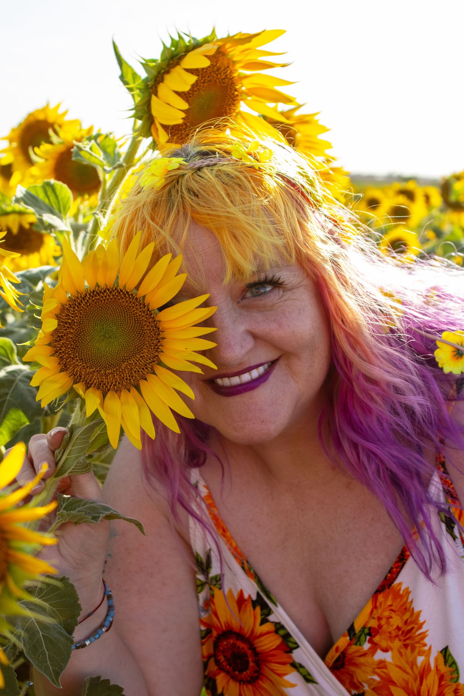 Smiling woman with colorful hair holding sunflower over eye in field.
