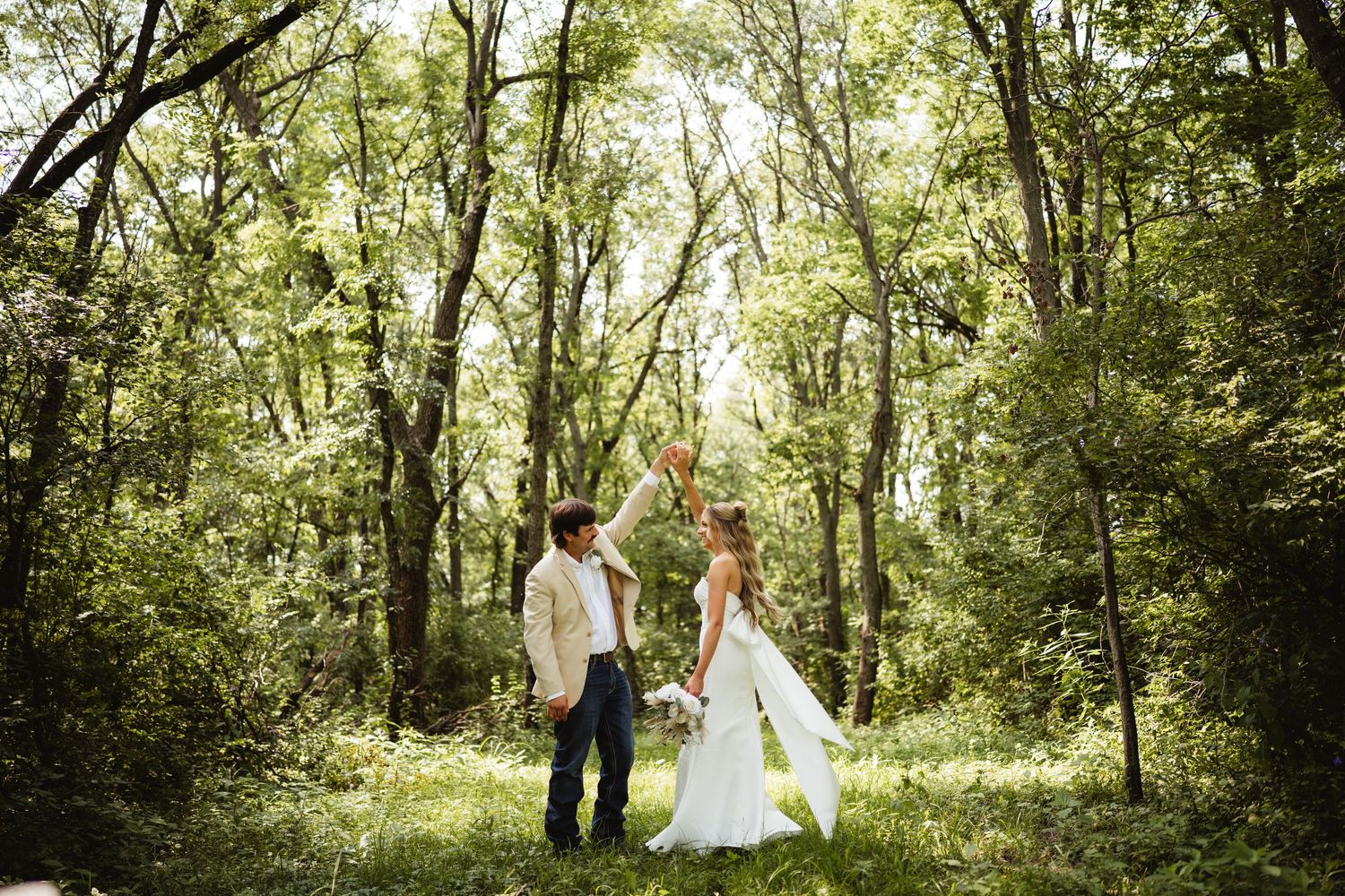 Bride and groom stand in lush green forest clearing, bride in white gown holding bouquet aloft on their wedding day.