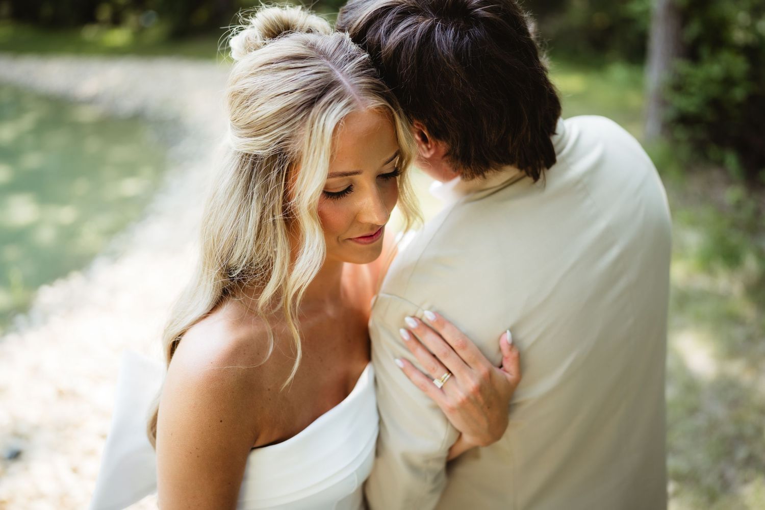Bride in white dress embraces groom in tan suit outdoors, showcasing her elegant half-up hairstyle and diamond ring.