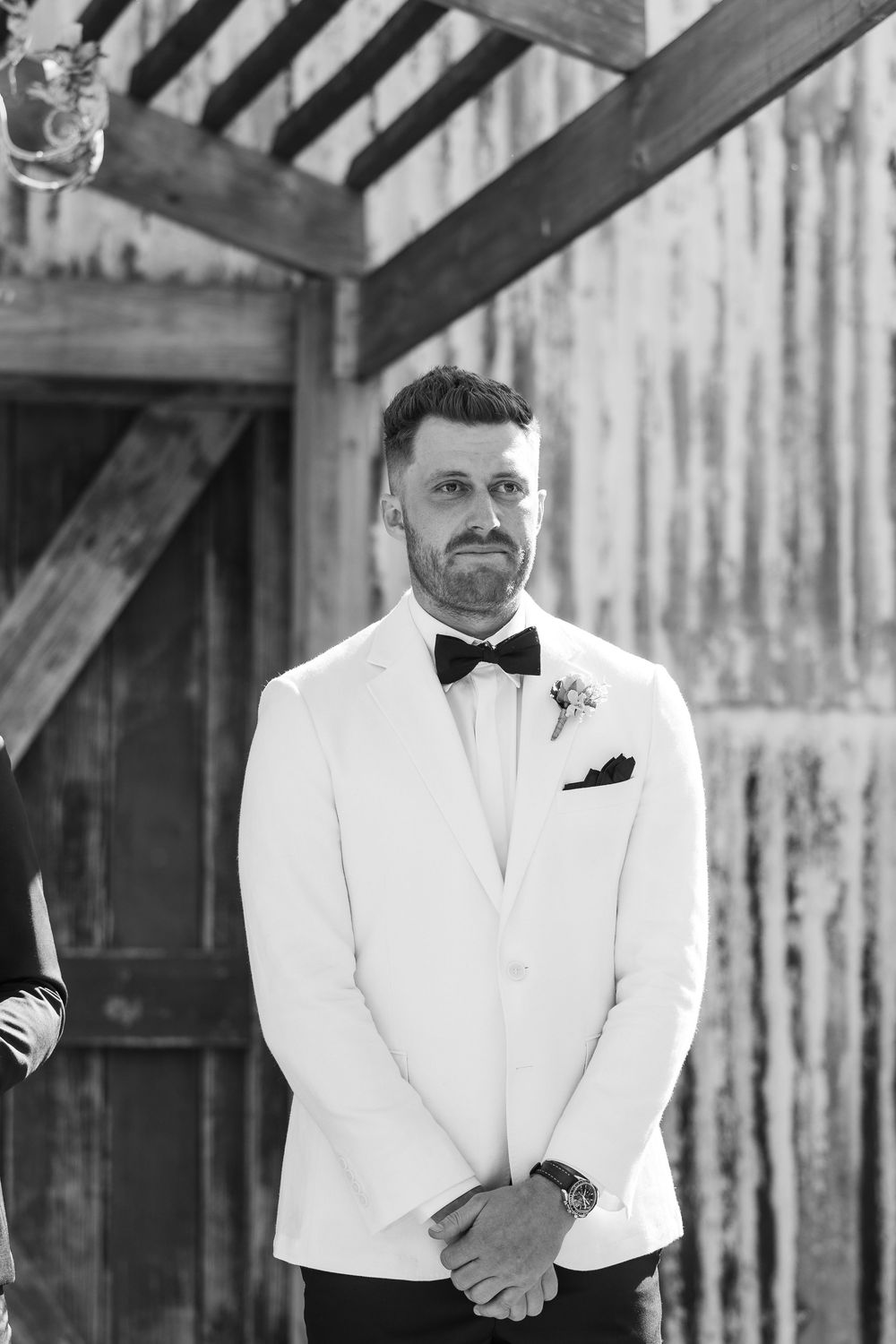 Black and white photo of a groom in white tuxedo jacket and bow tie, standing at the altar with emotional expression.