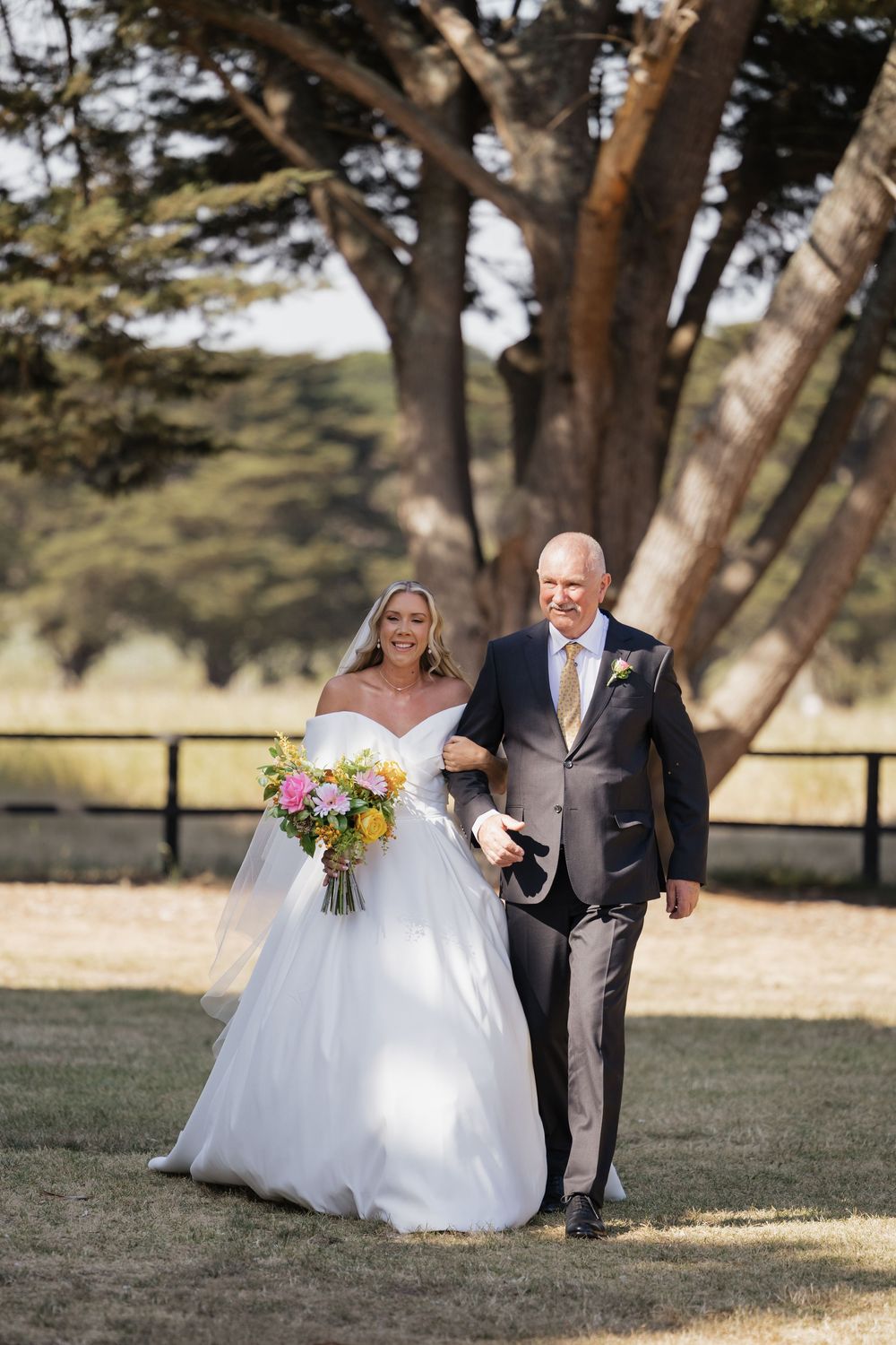 Bride in white off-shoulder gown holding colorful bouquet walks arm-in-arm with suited groom on sunny lawn.
