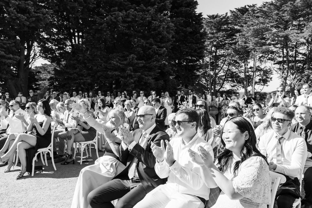 Black and white photo of a crowd clapping and smiling at an outdoor event surrounded by trees.