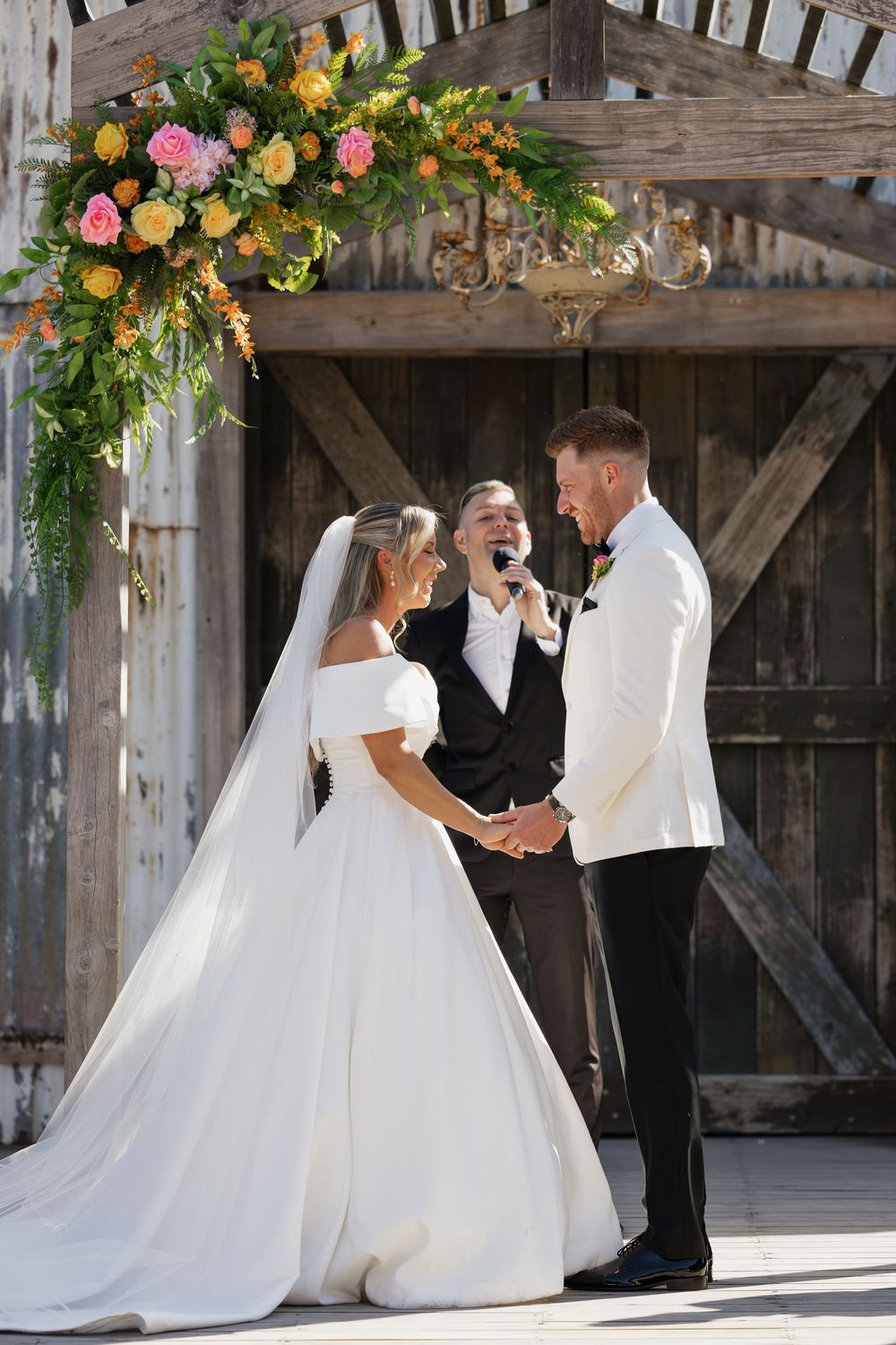 Bride and groom holding hands at outdoor barn wedding ceremony under colorful floral arch with officiant.