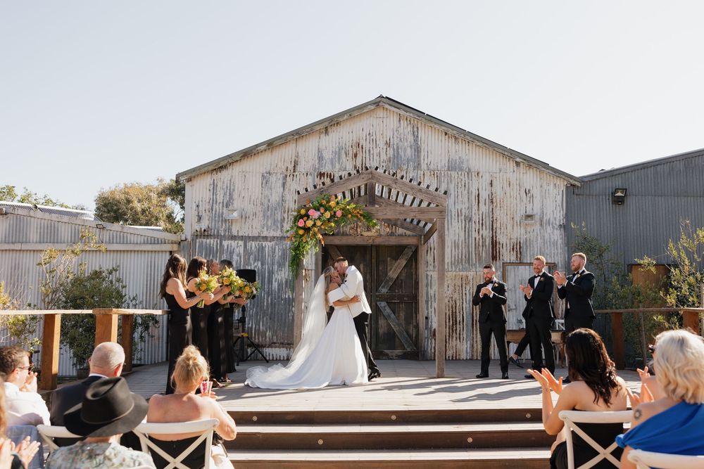 Couple shares first kiss at outdoor wedding ceremony in front of rustic barn with floral arch and seated guests.