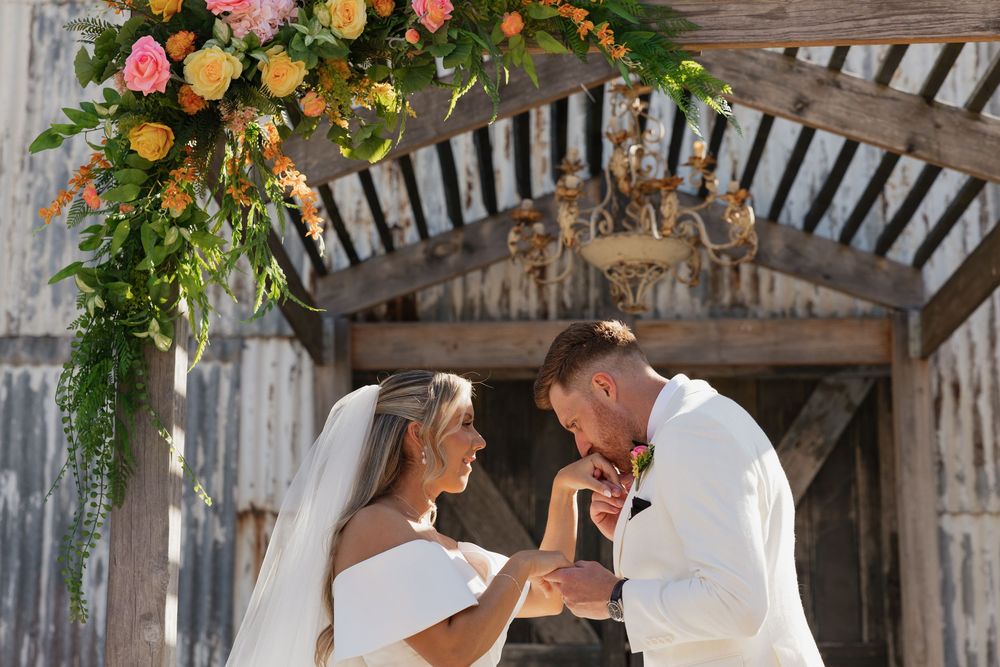Bride and groom exchanging rings under floral arch with pink and yellow flowers at rustic barn wedding ceremony.