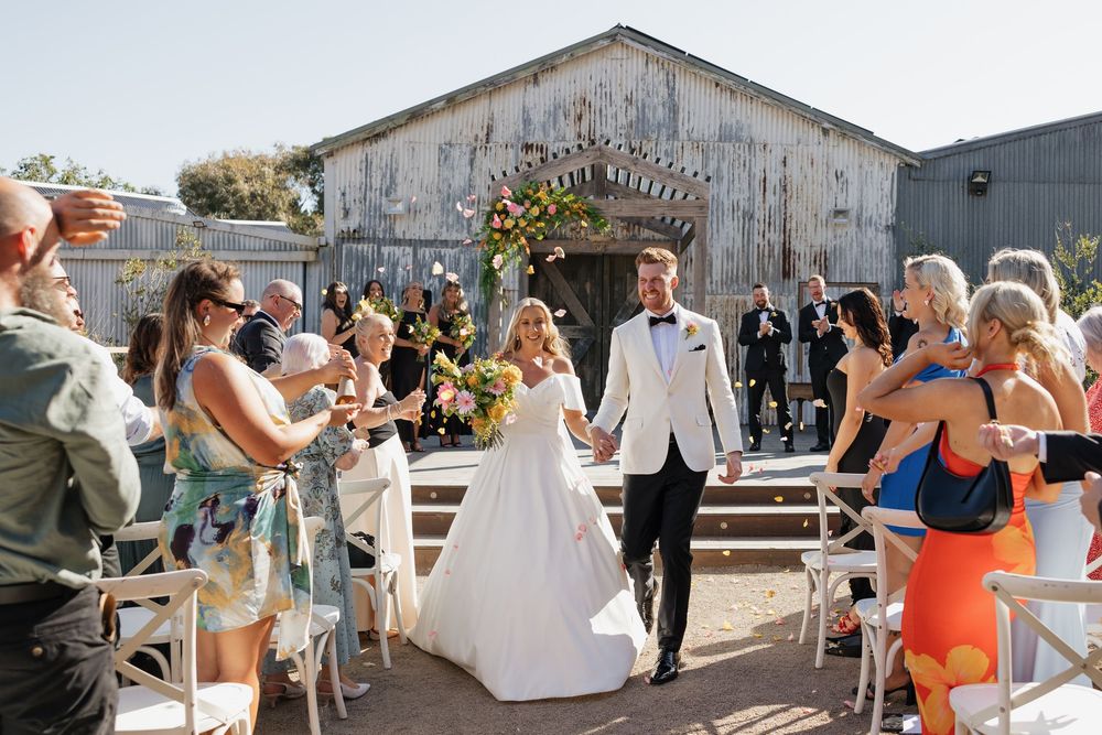 Bride and groom walk down outdoor aisle after wedding ceremony near rustic barn, guests celebrating around them.