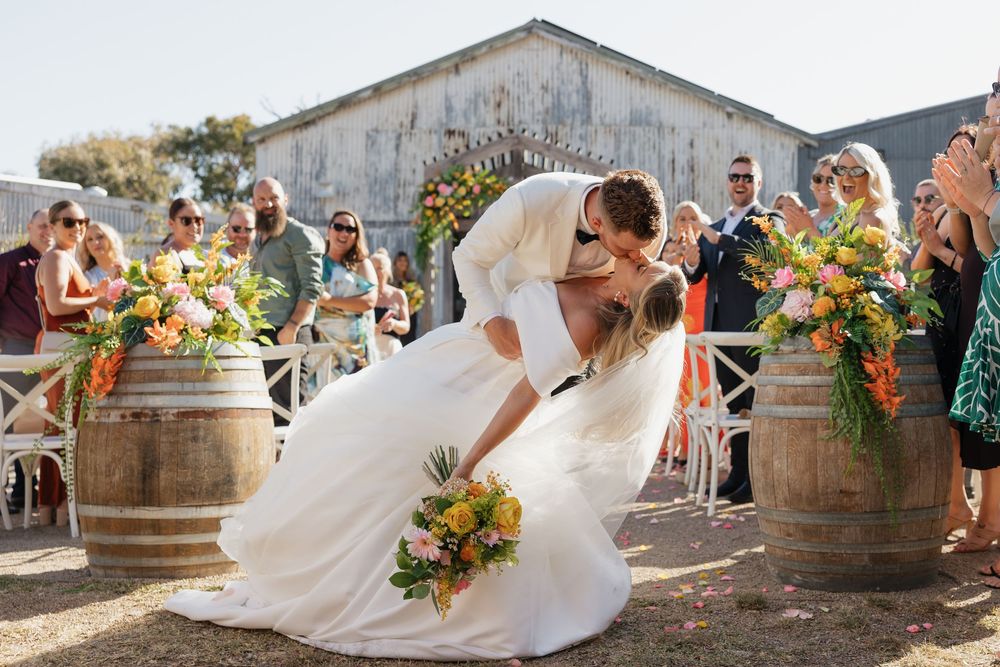Bride and groom share a romantic dip kiss at outdoor wedding ceremony flanked by wine barrels with vibrant tropical floral arrangements.