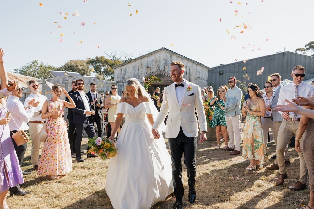 Bride and groom walk through confetti shower surrounded by smiling wedding guests outside rustic barn venue on sunny day.