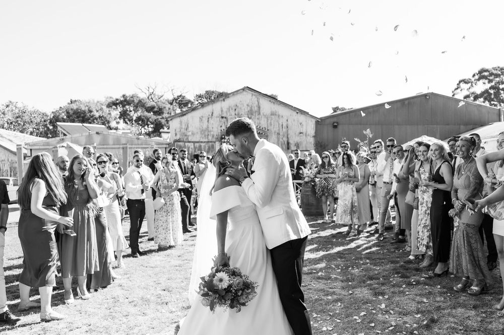 Black and white photo of bride and groom sharing a kiss while guests throw confetti during outdoor wedding ceremony.