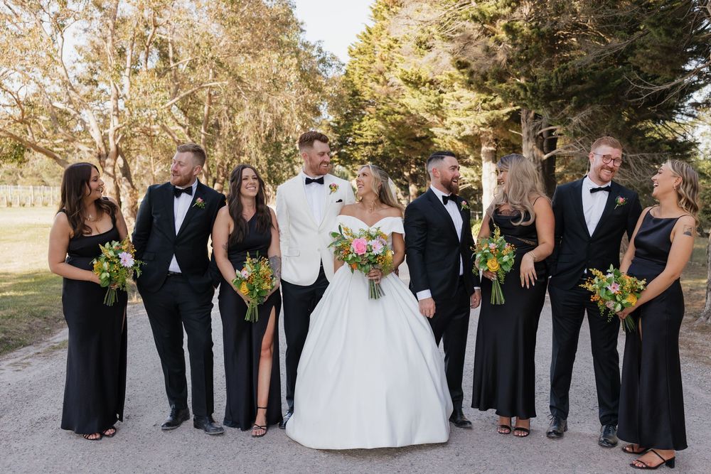 Wedding party in black attire with bride in white gown and groom in tuxedo posing outdoors among trees.