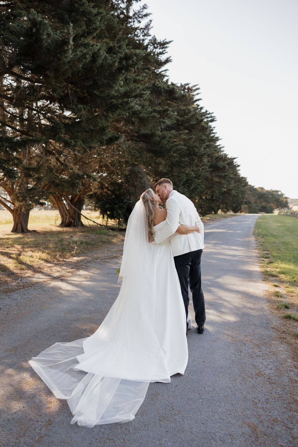 A bride and groom share a romantic kiss on a tree-lined path, the bride's long white veil trailing behind her.