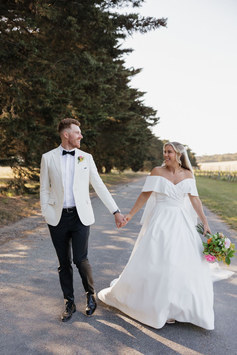 Bride and groom walk hand-in-hand down a tree-lined path, she in a white off-shoulder gown, he in a white tuxedo jacket.