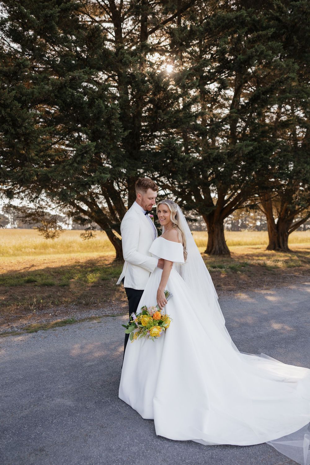Bride in off-shoulder gown with yellow bouquet and groom in white suit pose near trees at golden hour.
