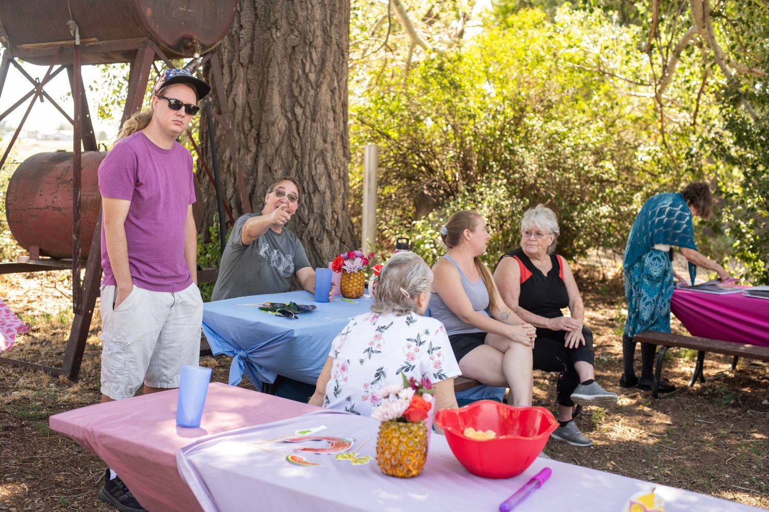 Group of people enjoying an outdoor picnic gathering under trees with fruit and drinks on a pink tablecloth.