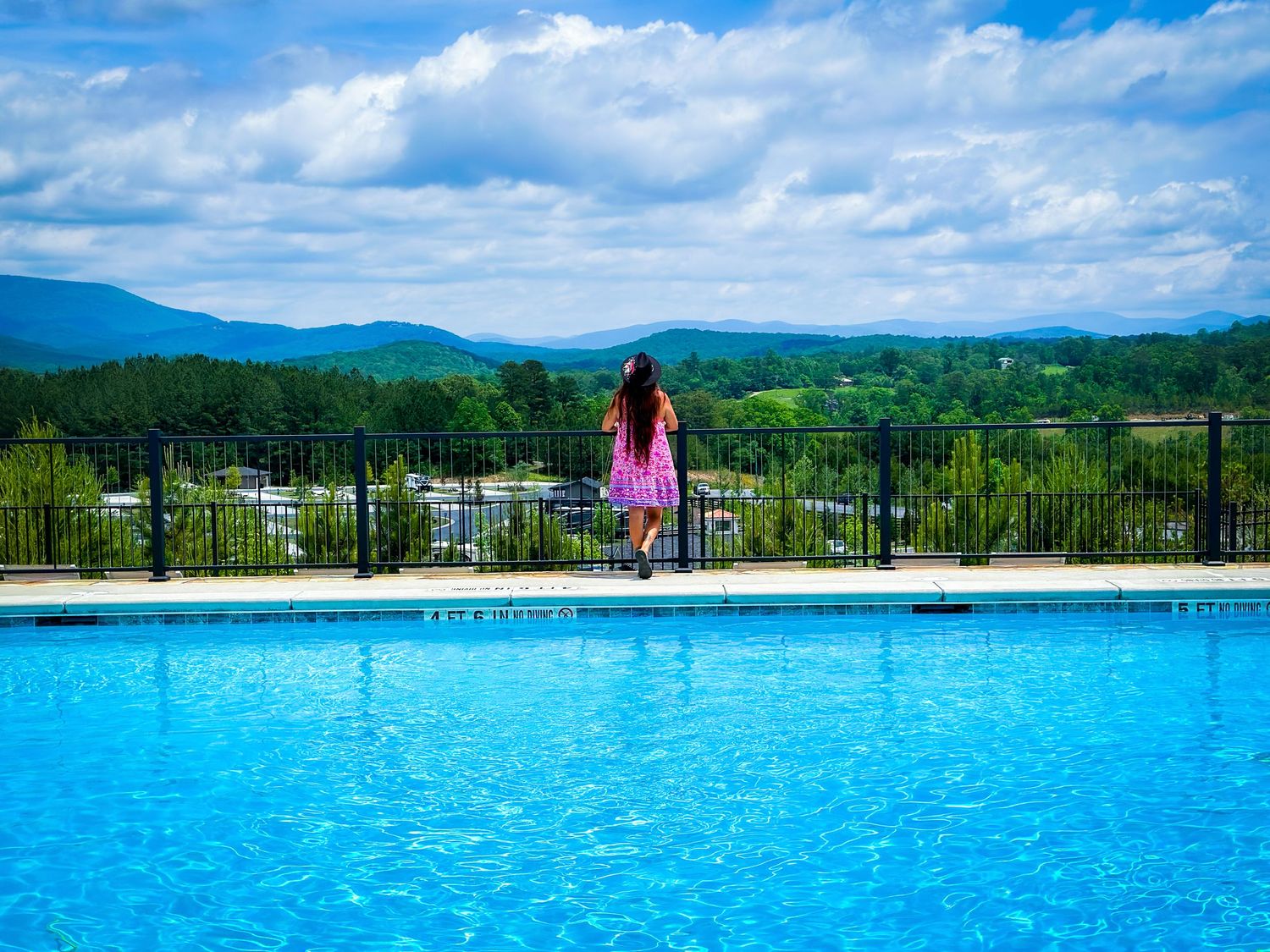 Woman in pink dress standing by infinity pool overlooking lush green mountain valley landscape.