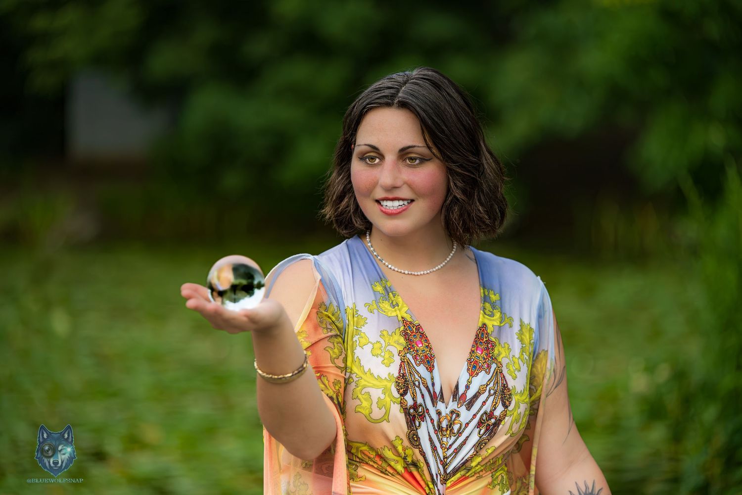 Woman in colorful floral dress holds crystal ball outdoors with green foliage background. Chi.Shooters meet gabbsbosh