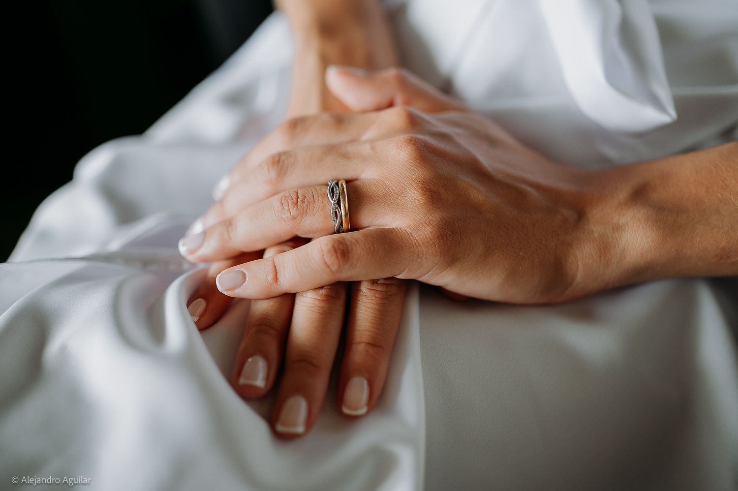 Close-up of two hands resting together on white satin, one wearing an elegant diamond engagement ring.