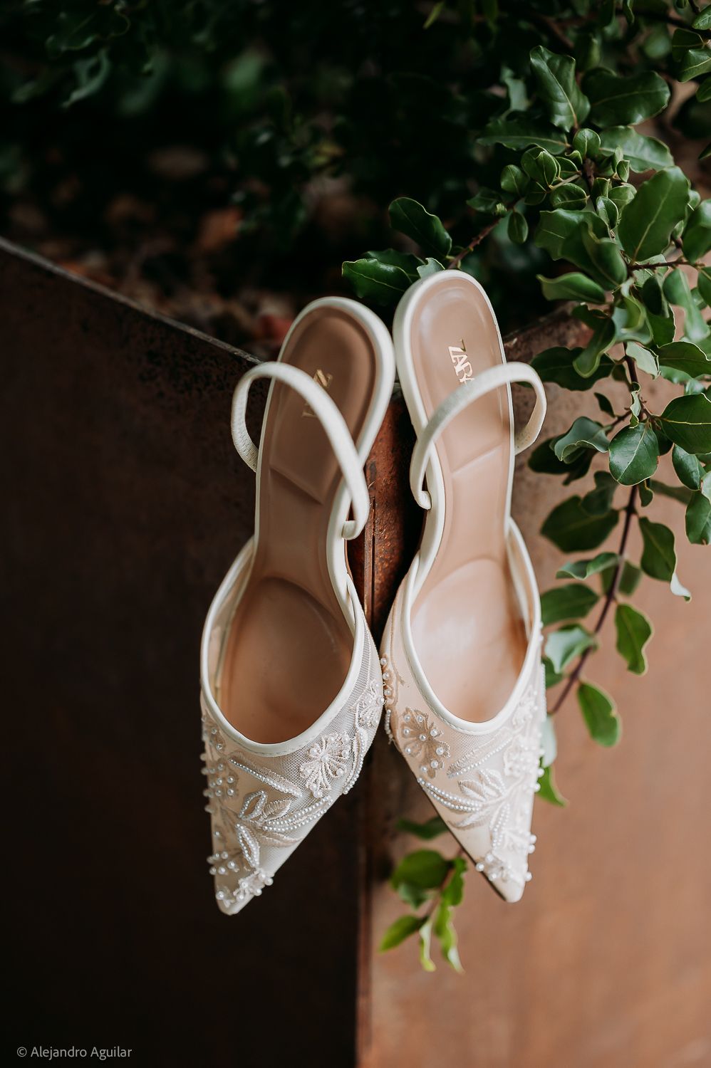 White beaded bridal heels with ankle straps and floral embroidery displayed against a rustic wall with greenery.