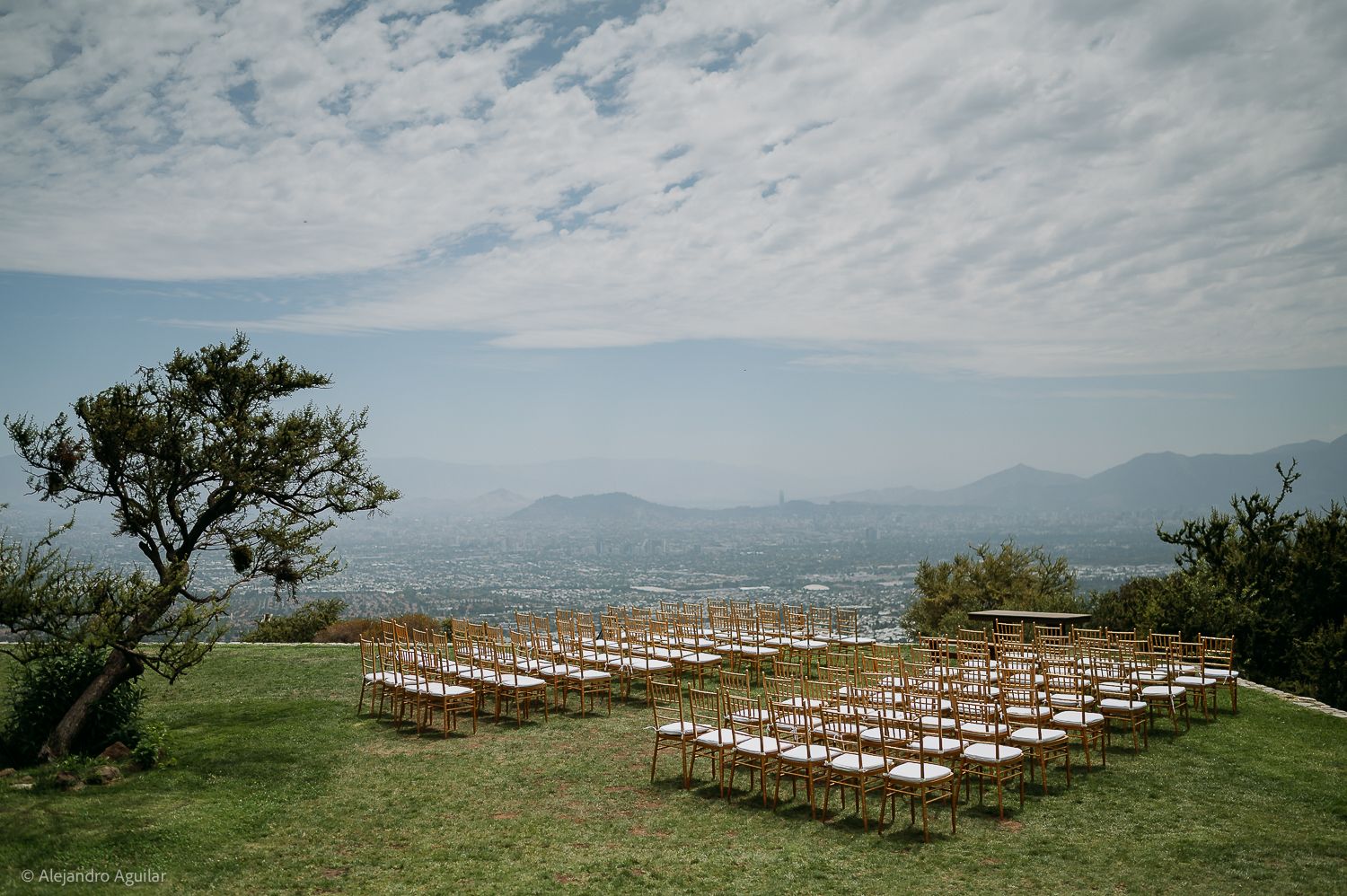 Outdoor wedding ceremony chairs arranged on a hilltop with panoramic mountain and city views under a cloudy sky.