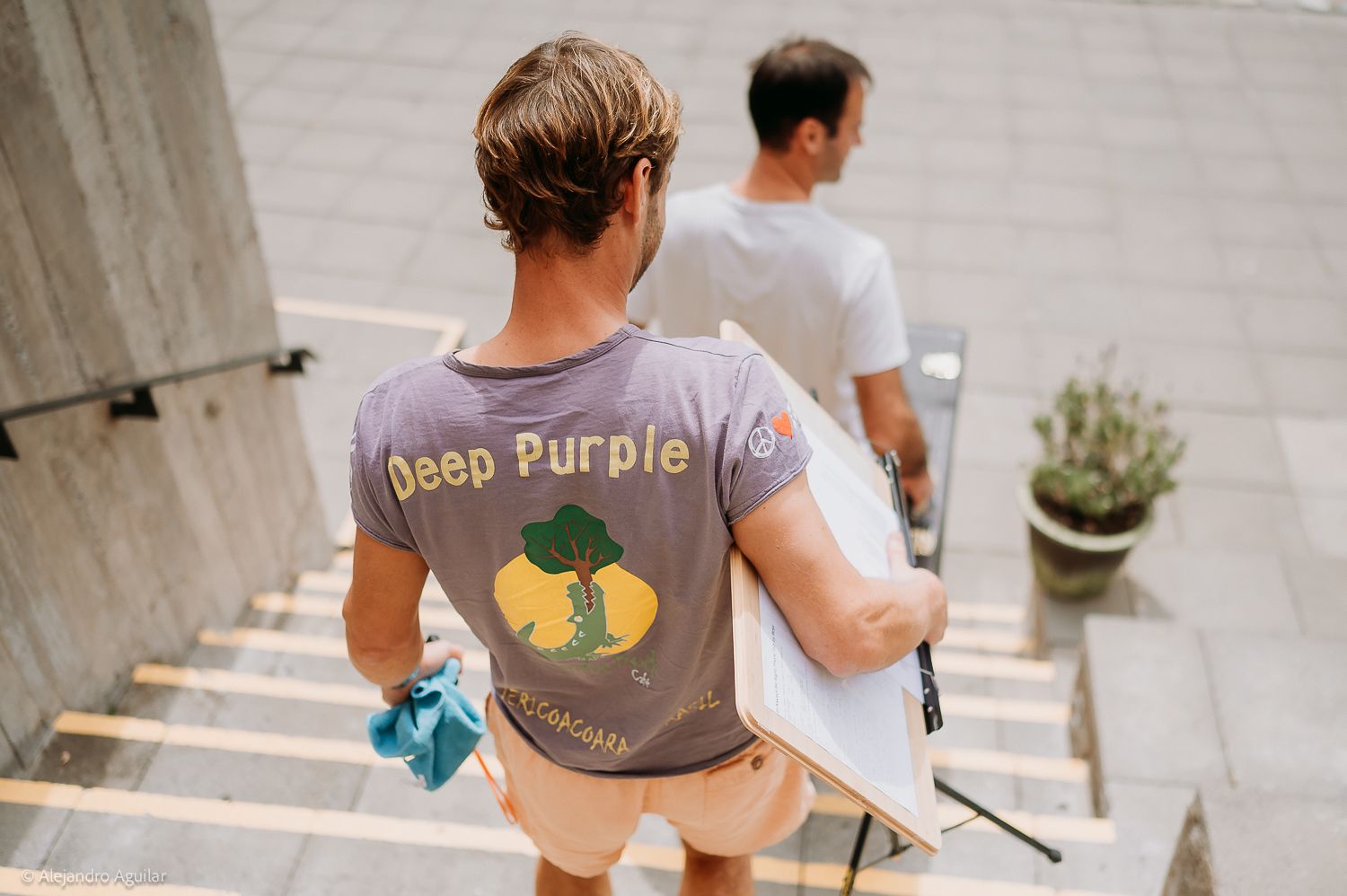 Two men walking up stairs, one wearing a gray Deep Purple graphic t-shirt, carrying boards outdoors.