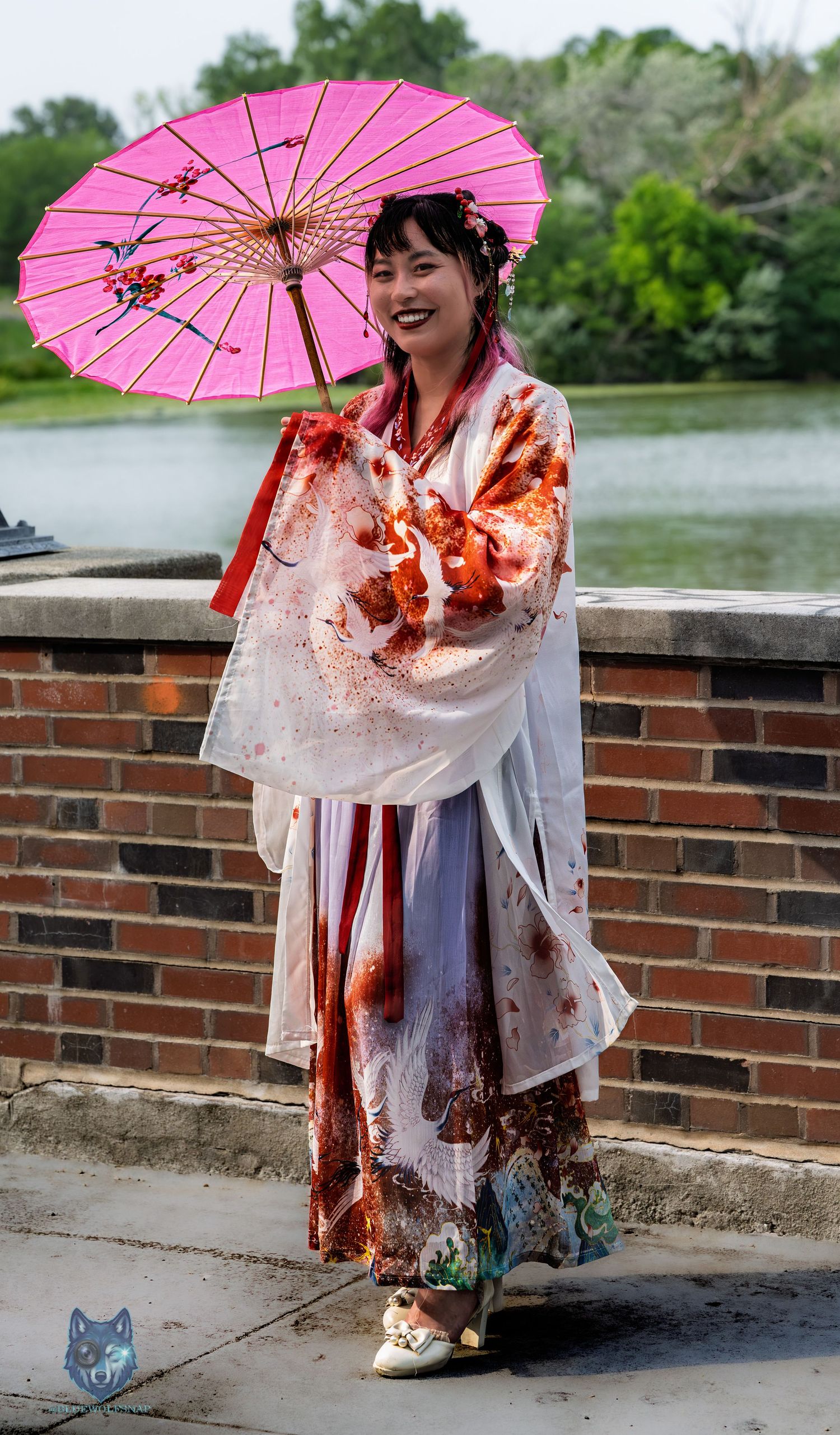Smiling woman in ornate white and red Hanfu cosplay holds pink floral parasol outdoors. Chi.Shooters meet yutonew