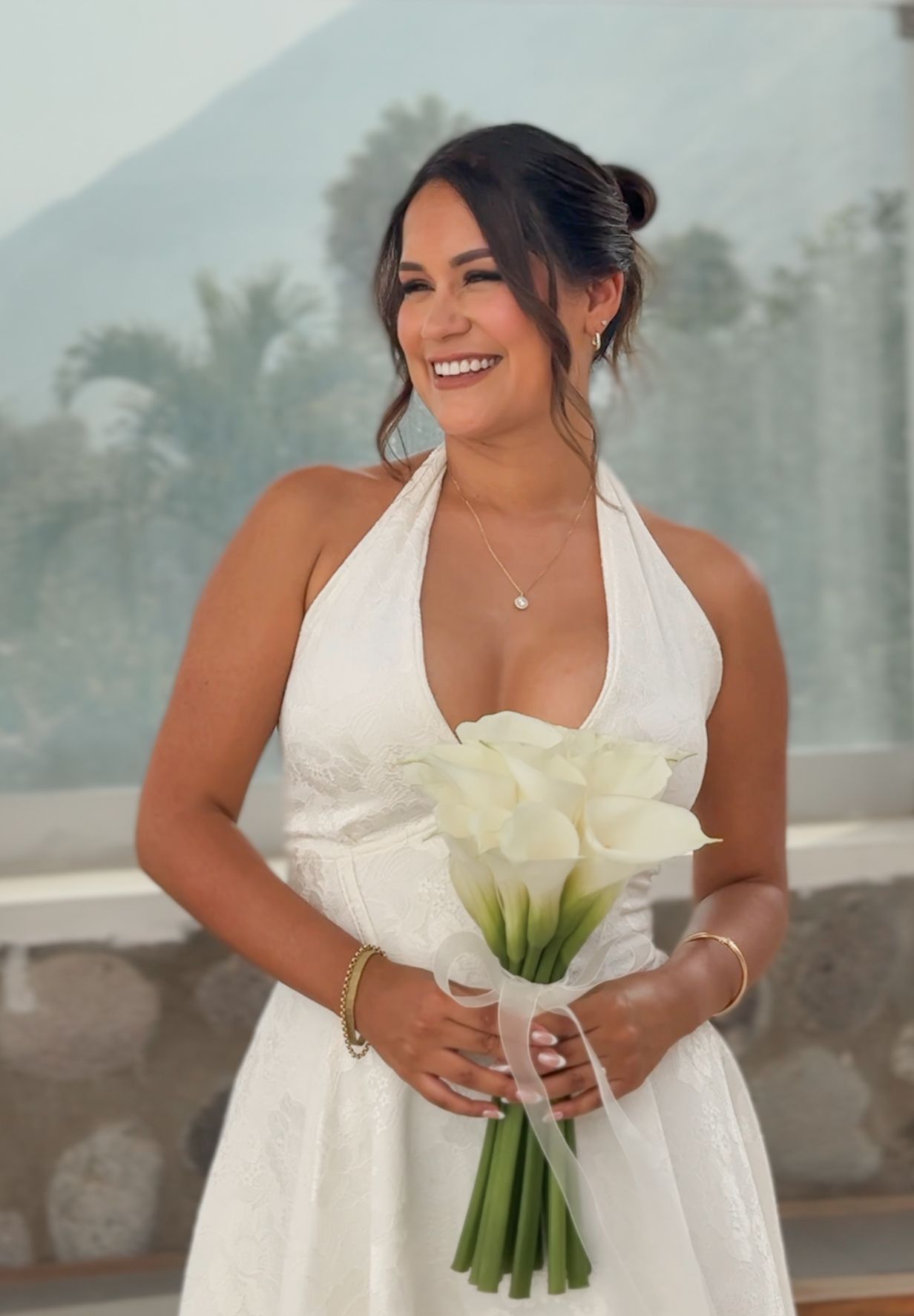Smiling bride in white halter wedding dress holding white calla lily bouquet with mountains and palm trees in background.
