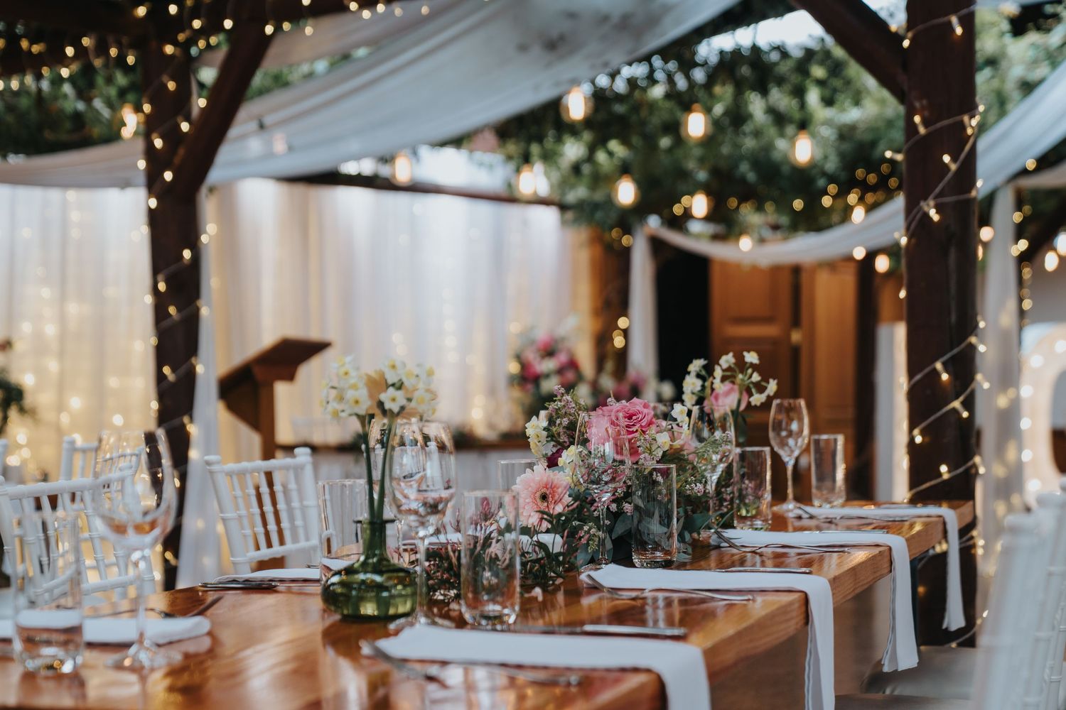 Elegant outdoor wedding reception with fairy lights, floral centerpieces, and wooden tables set for dining.