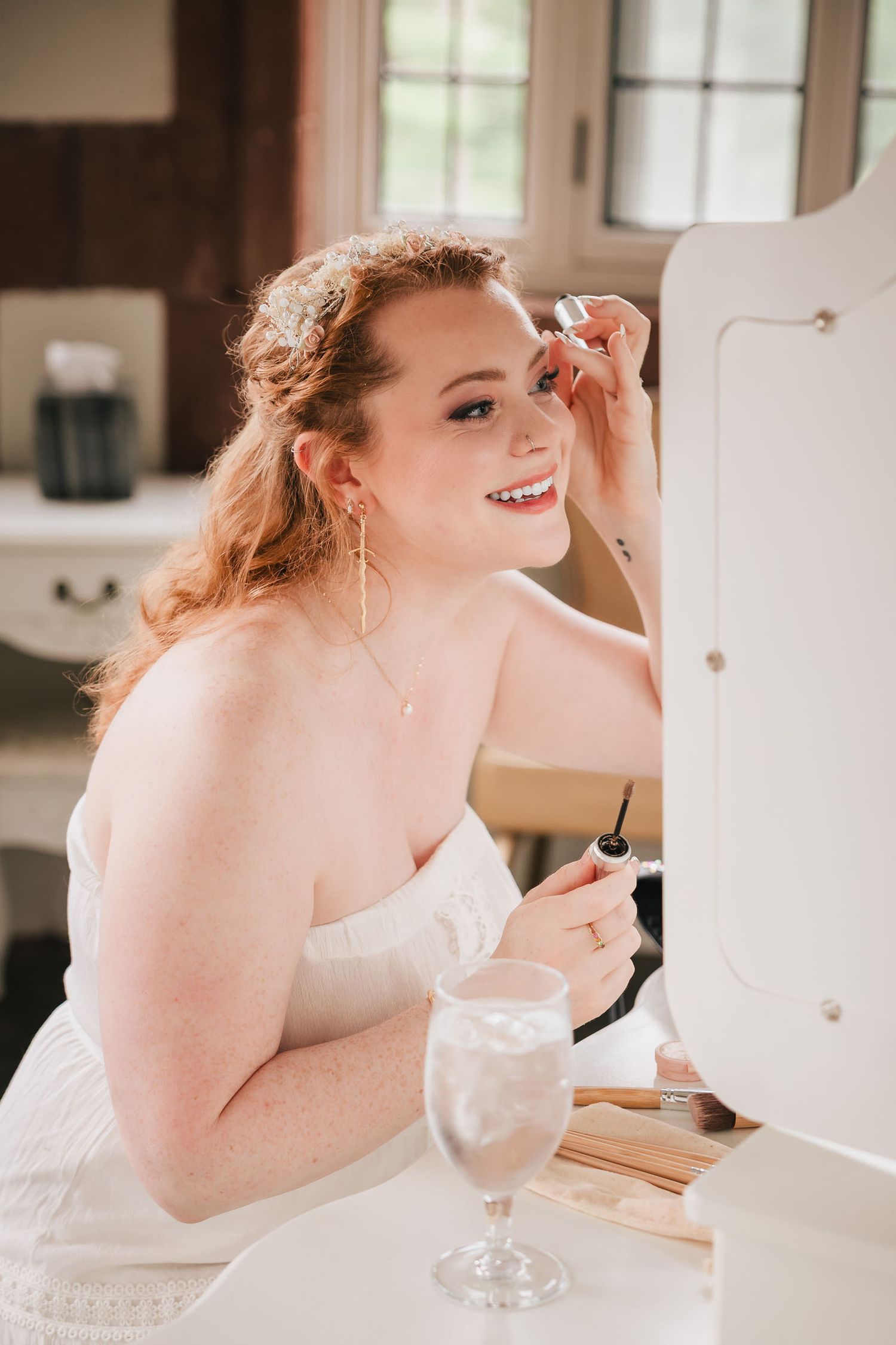 Bride applying her own mascara and smiling at her reflection during wedding day preparations.