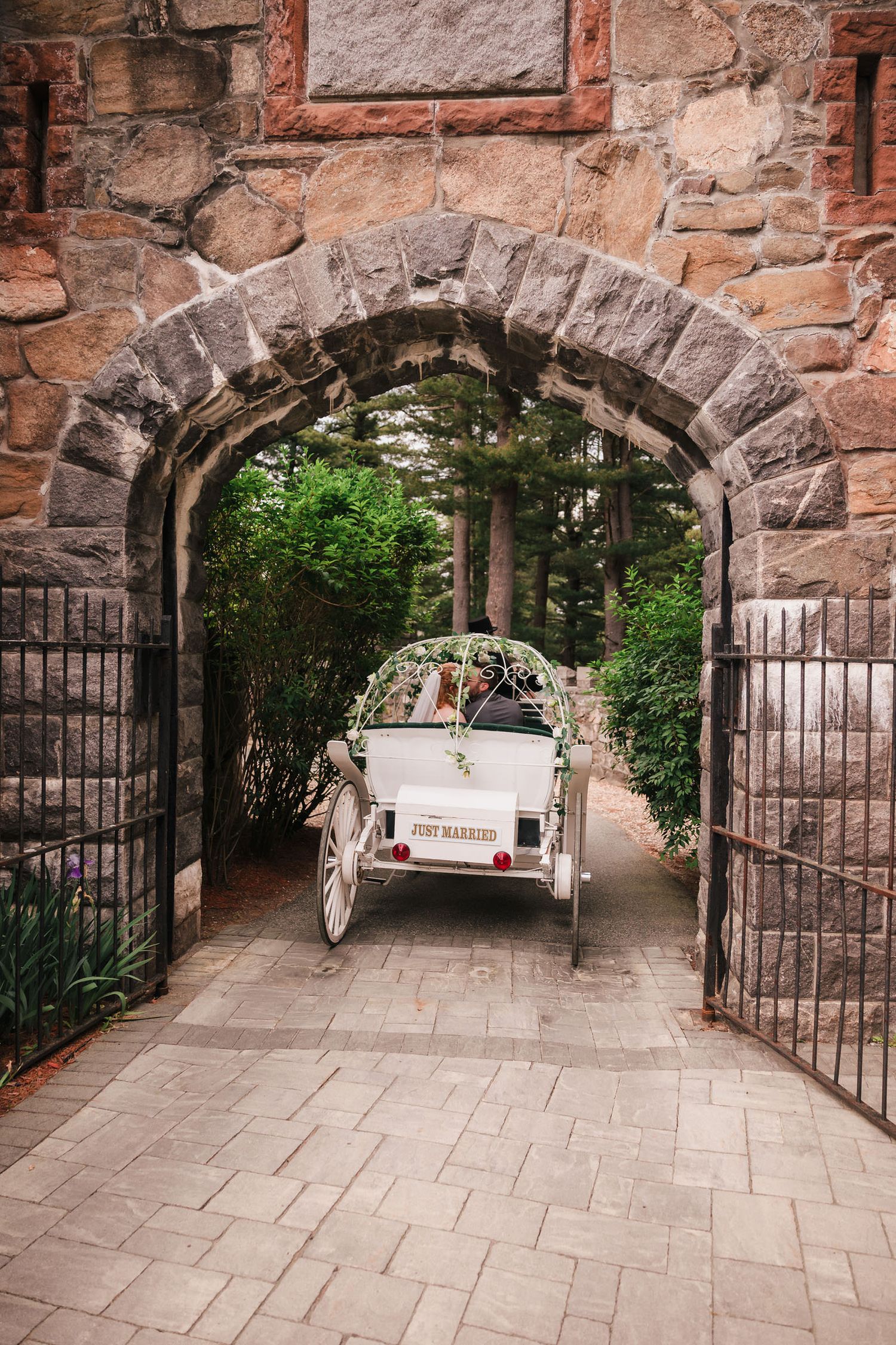 A 'Just Married' carriage carrying the newlyweds away through a stone archway lined with greenery.