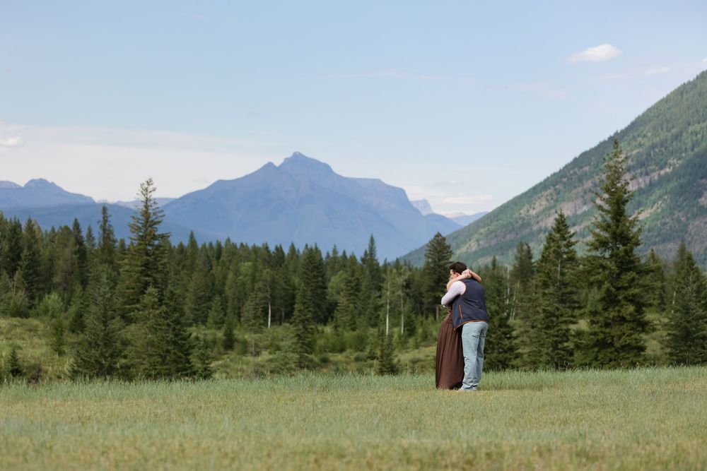 Couple embracing in green meadow with stunning mountain peaks and pine forest under clear blue sky.
