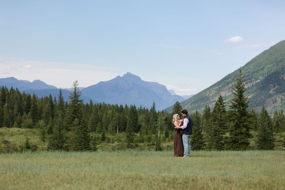 Couple embracing in a green meadow with tall pine trees and majestic mountain peaks under a clear blue sky.