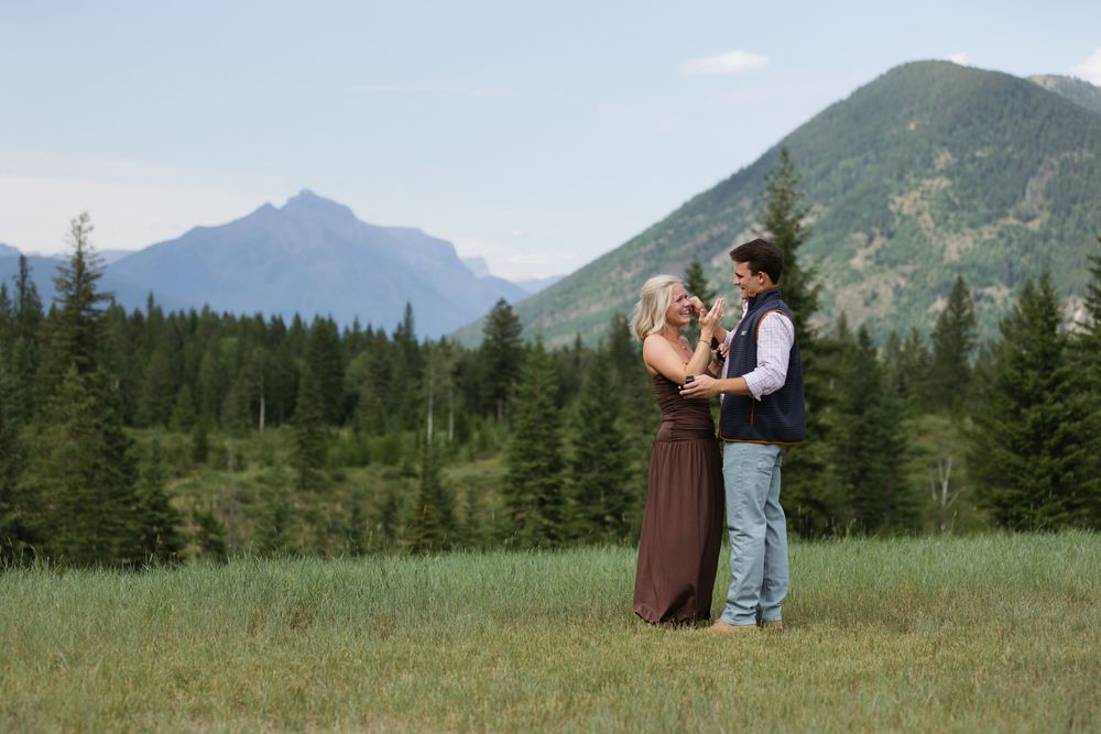 Couple embracing in a green meadow with pine trees and mountain peaks in the background during an outdoor engagement session.