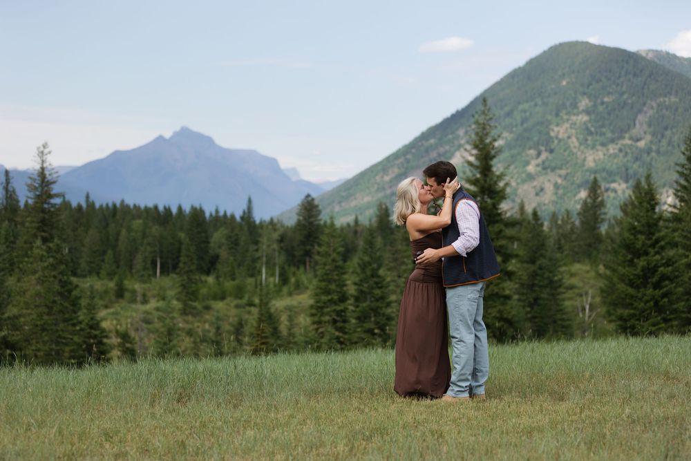 Couple embracing in a green meadow with pine trees and mountain peaks under a clear blue sky.