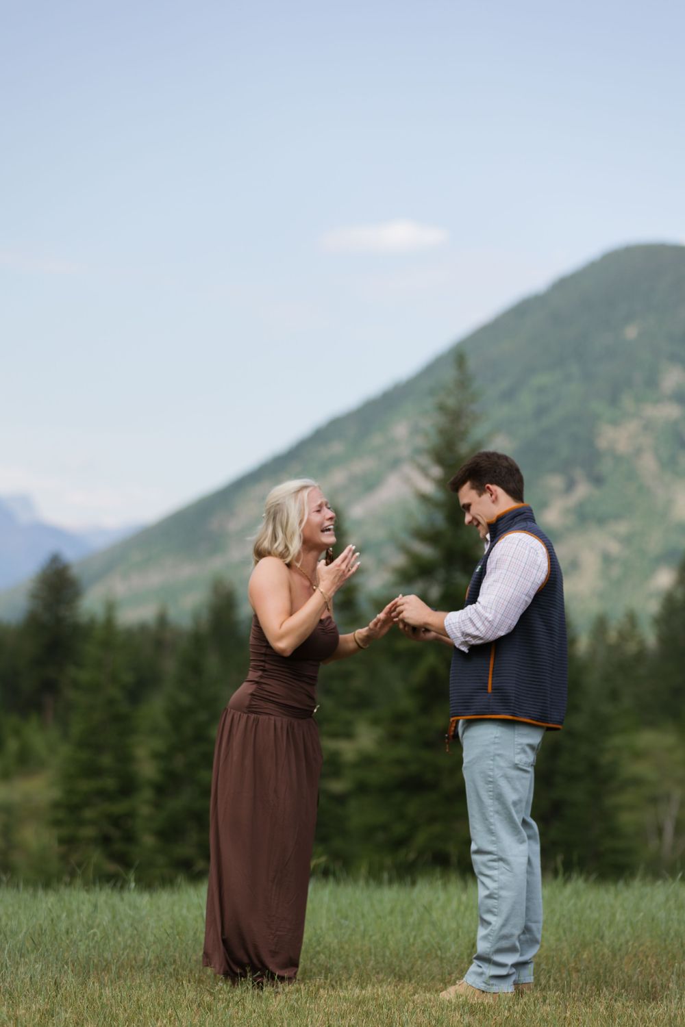Happy couple celebrating an engagement in a mountain meadow, woman in brown dress reacting with joy as man holds her hands.