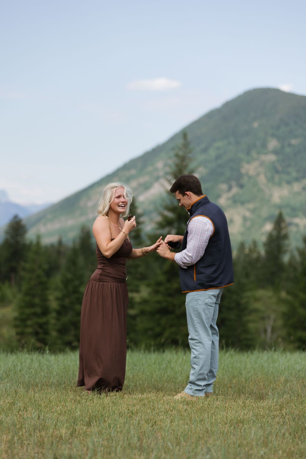 Couple sharing a joyful moment in a mountain meadow, man presenting ring to laughing woman in brown dress.