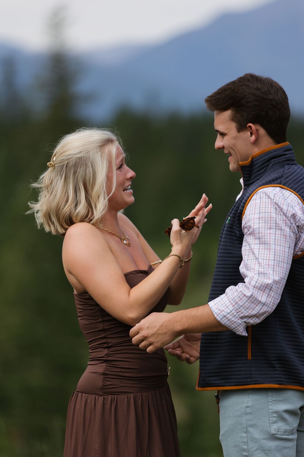 Couple sharing a joyful moment outdoors, woman in brown dress laughing while holding hands with man in plaid shirt and vest.