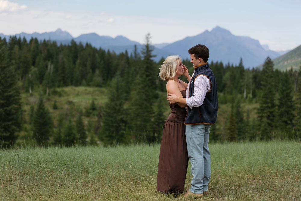 Couple embracing in a mountain meadow with evergreen forest and rocky peaks in the background.