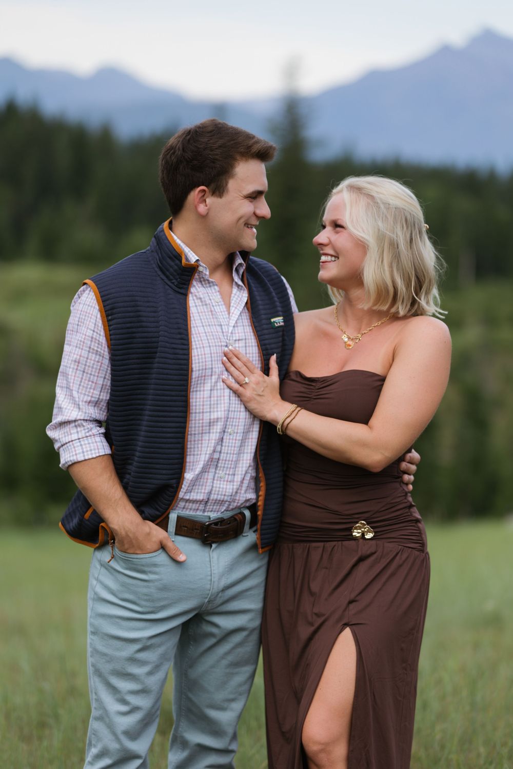 A smiling couple shares a joyful moment in a mountain meadow, the woman in a brown dress and man in a plaid shirt and vest.
